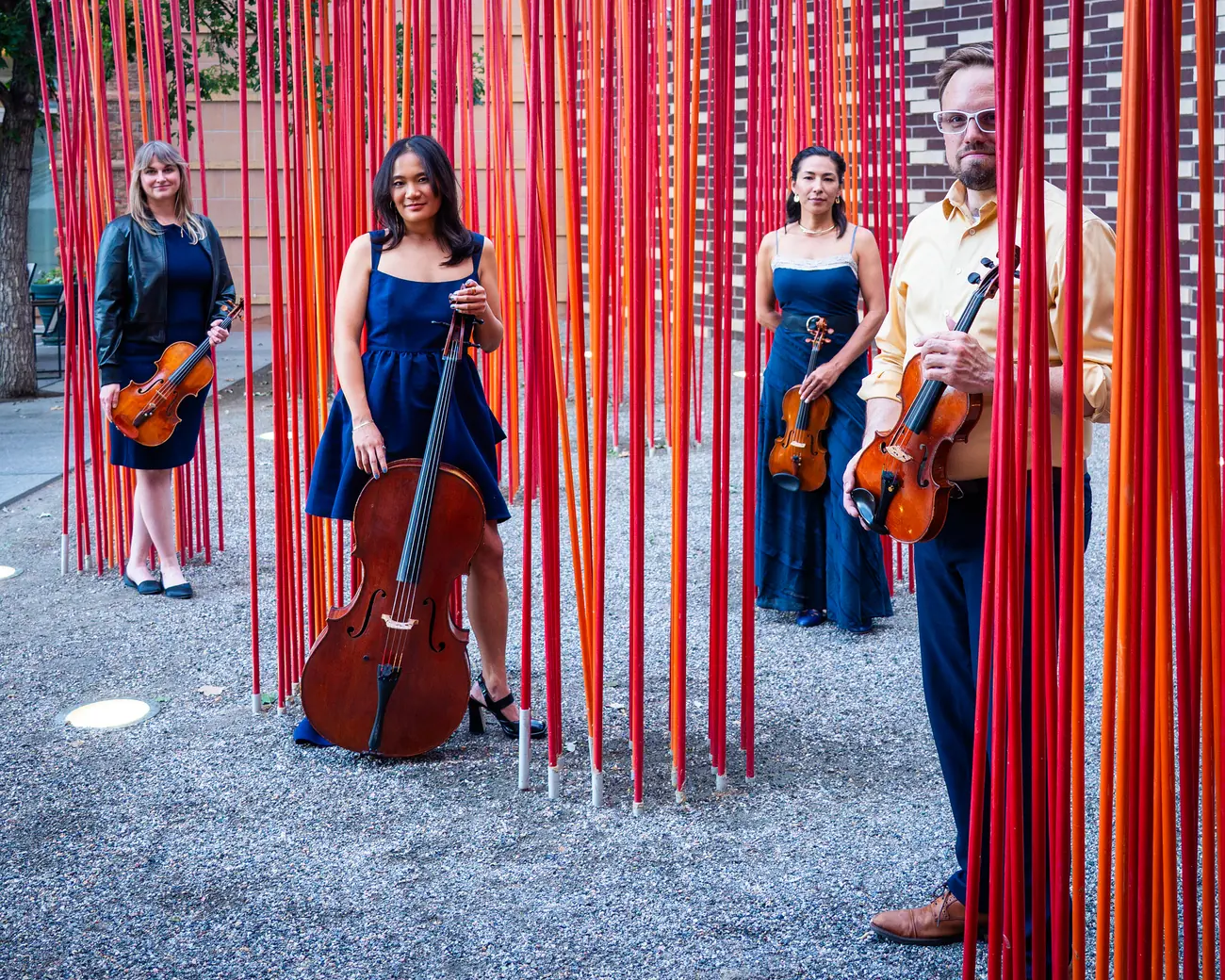 The quartet standing among a sculpture installation of tall vertical rods, photographed among red and orange rods